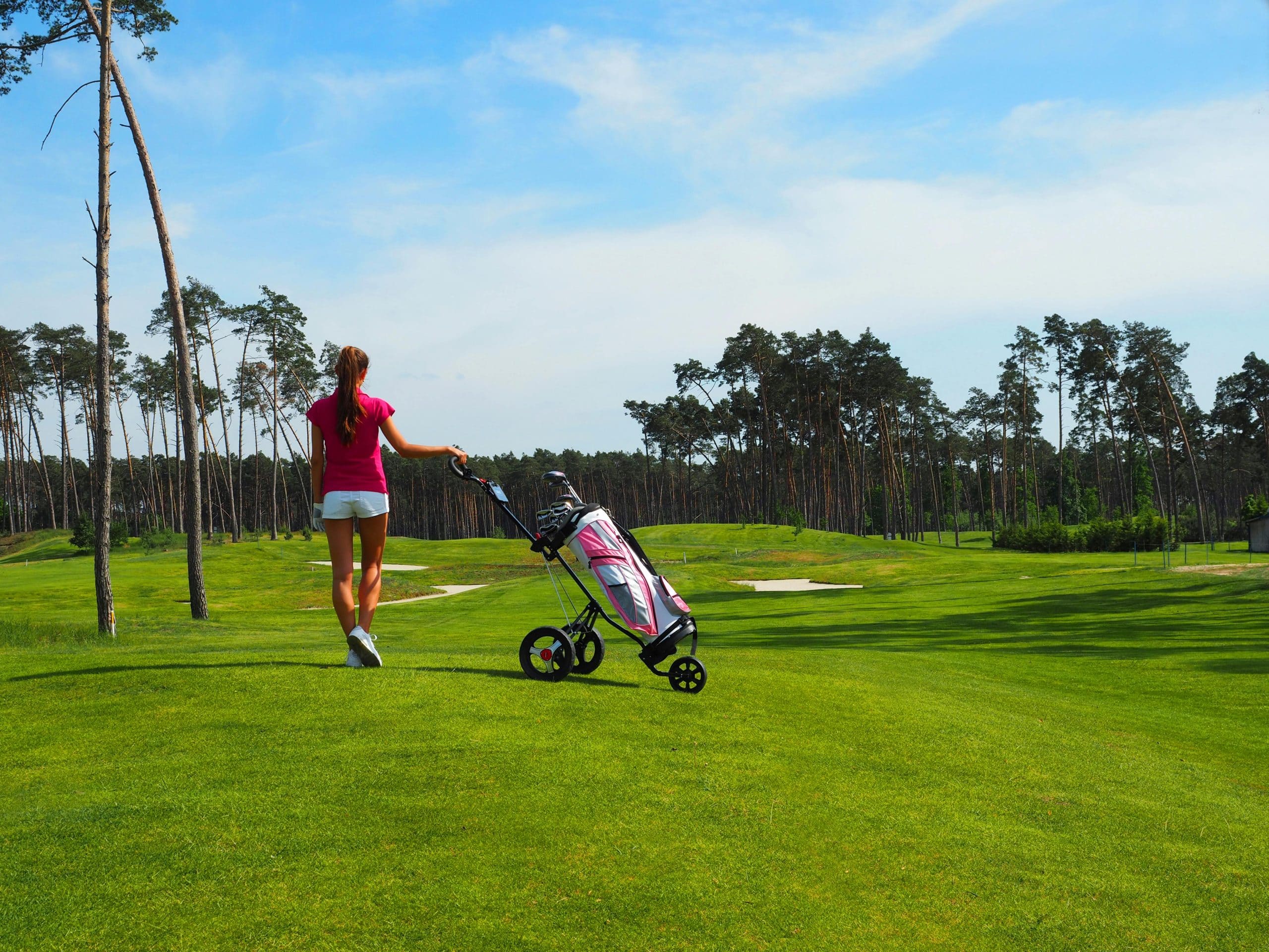 Femme poussant un chariot de golf sur un parcours verdoyant, ciel bleu.